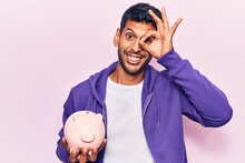 Young Latin Man Holding Piggy Bank Smiling Happy Doing Ok Sign With Hand On Eye Looking Through Fingers