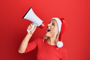 Wall Mural - Young blonde woman wearing christmas hat screaming using megaphone over isolated red background.