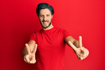 Young hispanic man wearing casual red t shirt smiling with tongue out showing fingers of both hands doing victory sign. number two.