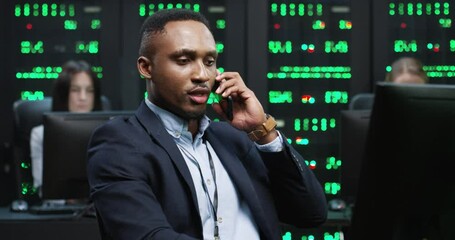Young handsome African American programmer sitting in office at workplace on background of working servers looking at computer and taking phone call on smartphone.