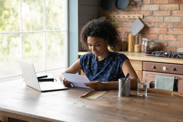 Satisfied African American woman reading letter, good news, money refund, businesswoman working with correspondence, sitting at table with laptop at home, received information from bank or college