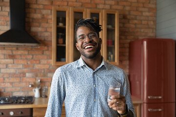 Wall Mural - Head shot portrait overjoyed African American man holding glass of fresh pure mineral water, standing in kitchen, looking at camera, healthy lifestyle and good daily habit concept, body and skin care