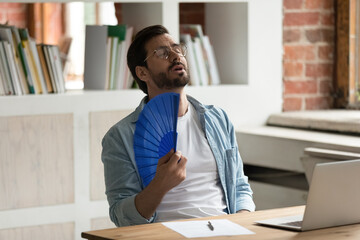 Unwell young Caucasian male employee sit on desk work on computer wave with hand fan. Overheated man worker use waver suffer from heatstroke in office, struggle with no AC at workplace.