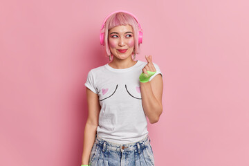 Pretty smiling girl with bob pink hair wears wireless headphones on ears makes korean like sign shapes mini heart with fingers listens favorite music dressed in t shirt and jeans poses indoor