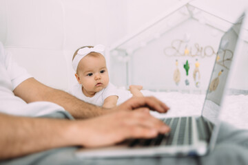 The young dad's hands are on the laptop keyboard, and the little daughter is looking at the screen. They are on the bed. The concept of remote work.