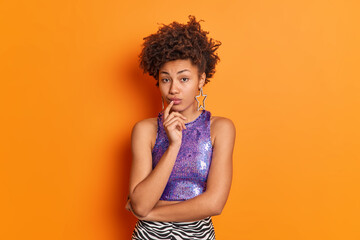 Wall Mural - Portrait of good looking serious African American woman looks directly at camera keeps finger on lips dressed in fashionable outfit poses at studio against orange background. People and style