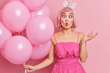 Beautiful young Asian woman makes fish lips has funny grimace raises hand wears bright makeup raises hand holds helium balloons isolated over pink background celebrates anniversary or birthday