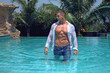 Muscular handsome sexy young wet man with athletic body is standing in the pool in blue jeans and white shirt. Fitness male model posing on camera