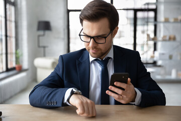 Canvas Print - Focused millennial Caucasian male employee in suit look at hand watch clock checking time meet deadline. Serious young businessman hold use modern smartphone, synchronize gadget with smartwatch.