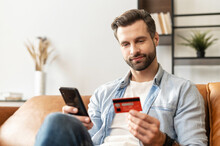 Bearded Handsome Man Makes Purchase Online. A Hipster Wearing Casual Shirt Holds A Mobile Phone And A Credit Card Orders Food. Smiling Guy Is Using Smartphone And Debit Bank Card For Paying In E-shop