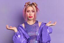Photo Of Clueless Young European Woman Looks Sadly Has Two Hair Buns Spreads Palms Faces Difficult Choice Wears Bright Vivid Makeup Dressed In Fashionable Clothes Isolated Over Purple Background