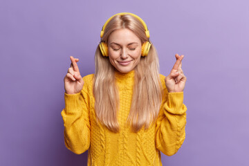Wall Mural - Hopeful blonde woman keeps eyes closed keeps fingers crossed makes desirable wish awaits for good news listens music via stereo headphones wears yellow sweater isolated over purple background