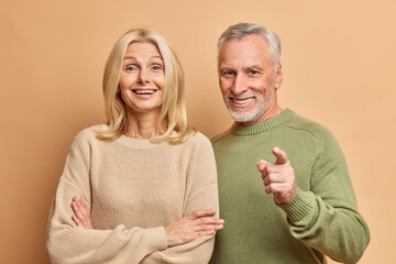 Sticker - Portrait of happy aged woman and man stand closely to each other dressed in casual jumpers look with great interest in front isolated over brown studio background. People age and emotions concept