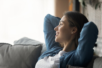 Captured by dreams. Happy serene millennial woman relax on sofa with hands over head look away dream imagine. Calm peaceful young lady sit in soft pillows rest alone remember good moments. Copy space