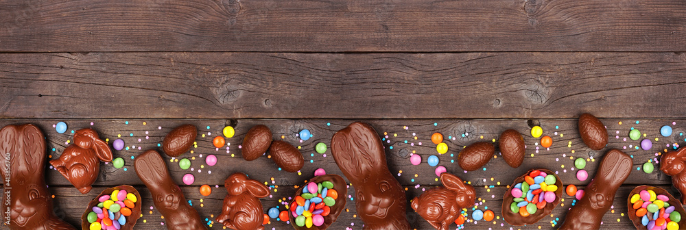 Easter candy and chocolate bunnies and eggs. Bottom border against a rustic dark wood banner background. Copy space. - obrazy, fototapety, plakaty 