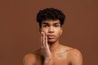 © Ivan - Photo of attractive black man with piercing touches his smooth face and looks at camera. Naked torso, isolated brown color background