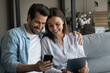 © fizkes - Millennial couple in love confident internet users sit on couch hold phone tablet exchange data share information between devices. Young husband wife send receive files app from cell to pad using wifi