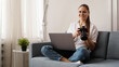 © brillianata - Happy young woman with photo camera using laptop at home. The photographer is holding the camera and smiling.