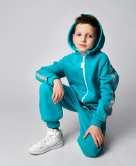 Fashion boy child in warm sportswear suit sits on one knee looking at camera. Male child advertises sports outerwear. Sporty fashionable outfit. Studio portrait