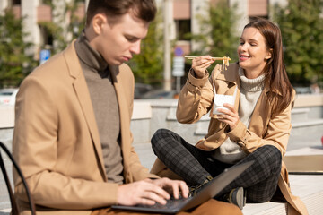 Poster - Young serious elegant man using laptop while his happy girlfriend eating wok