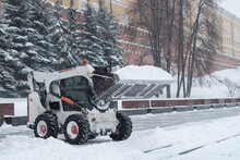 A Small Loader Excavator Bobcat Removes Snow From The Sidewalk Near The Kremlin Walls During A Heavy Snowfall. Snowflakes Are Flying In The Air.
