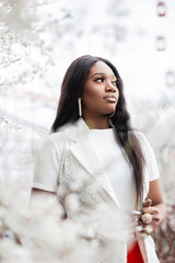 Wall Mural - Fashionable beautiful young black woman in stylish white clothes walks near a flowering tree in nature. Stylish african girl with attractive appearance outdoors