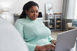 © Seventyfour - Portrait of curvy African American woman using laptop while enjoying work from home relaxing on couch in modern minimal interior