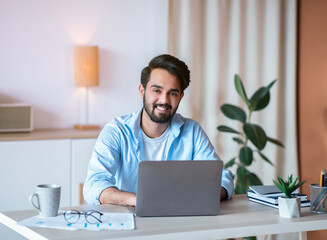 Wall Mural - Remote Career. Happy Arab Male Entrepreneur Sitting At Desk With Laptop