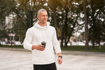 Man dressed white hoodie with coffee in hand waiting for someone in summer park Caucasian guy standing outdoors smiling