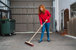 Smiling female worker sweeping the warehouse with a large broom