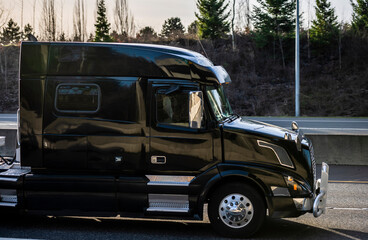 Black industrial shiny big rig semi truck with grille guard and sleeping compartment in the cab transporting cargo driving on the highway road