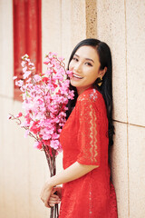 Sticker - Happy beautiful young Vietnamese woman in red dress standing outdoors with brahches of pink peach blooming branches