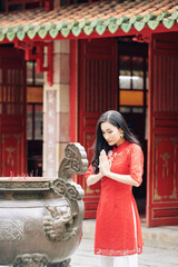 Poster - Pretty calm young Vietnamese woman in red dress praying at ancient bronze urn with incense sticks at Buddhist temple