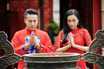 Poster - Young couple standing with eyes closed and holding hands in pray gesture when standing at ancient bronze urn with incense sticks