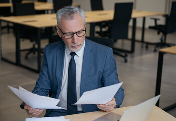 Pensive senior businessman reading financial report, analyzing business contract, brainstorming. Portrait of handsome banker working with paper documents sitting in office