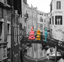 Venice Carnival. Three Masks In Checkered Colorful Costumes Standing On The Bridge Over Canal. Retro Toned Black White Selective Color Photo.
