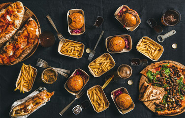 Lockdown takeaway fast food dinner from delivery service concept. Flat-lay of quarantine home party with burgers, french fries, sandwiches, pizza, beer in glasses over dark table, top view