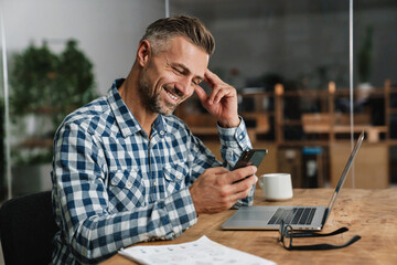 smiling grey man using mobile phone while working with laptop