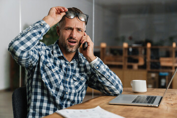 Wall Mural - Shocked grey man talking on mobile phone while working with laptop