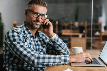 Wall Mural - Pleased grey man talking on mobile phone while working with laptop
