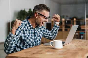 Wall Mural - Excited man making winner gesture while working with laptop