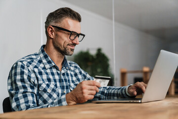 Wall Mural - Pleased unshaven man using credit card while working with laptop
