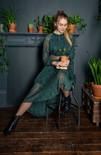 A Blonde Girl In A Green Dress With A Book In Hand In A Cozy Room.