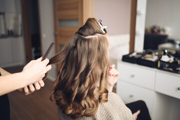 Wall Mural - Hair stylist prepares beautiful young woman for event, makes curls hairstyle with a curling iron for client in beauty salon. Long beautiful light brown natural hair.