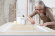 © amedeoemaja - wood crafts, woman artisan carpenter painting with brush and paint jar white the door in workshop, wearing overall and eyeglasses, interior designer, restoration, diy and handmade works concept