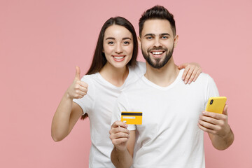 Wall Mural - Young couple two friends man woman in white basic blank print design tshirts hold in hand using mobile cell phone credit bank card order home delivery in restaurant isolated on pastel pink background