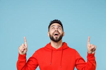 Wall Mural - Young caucasian smiling bearded attractive man in casual red orange hoodie point index finger overhead on copy space area mock up isolated on blue background studio portrait People lifestyle concept.