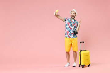 Wall Mural - Full length of cheerful young traveler tourist man in hat doing selfie shot on mobile phone showing victory sign isolated on pink background. Passenger travel on weekends. Air flight journey concept.