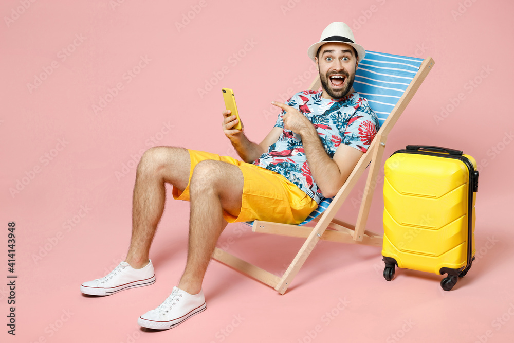 Full length excited young traveler tourist man in hat sit on deck chair pointing index finger on mobile cell phone isolated on pink background. Passenger travel on weekend. Air flight journey concept. - obrazy, fototapety, plakaty 