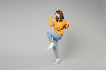 Full length of young smiling positive overjoyed caucasian happy satisfied woman 20s in knitted yellow sweater do winner gesture clench fist looking camera isolated on grey background studio portrait